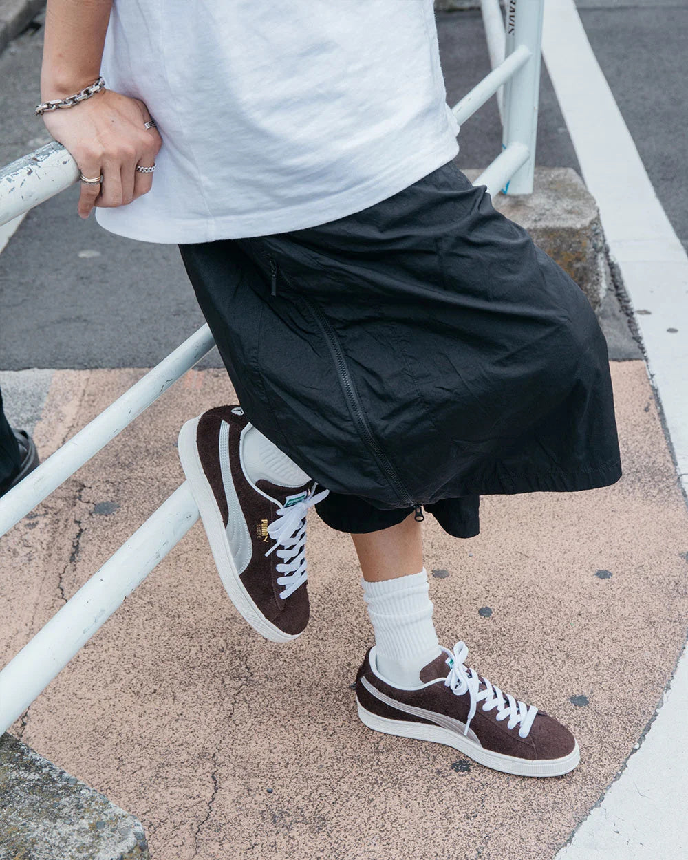 On-foot shot of the BILLY'S ENT x PUMA Suede Classic Chocolate Fondue in shaggy brown suede with a silver formstrip and white laces, leaning on a street railing. Photo via Fullress