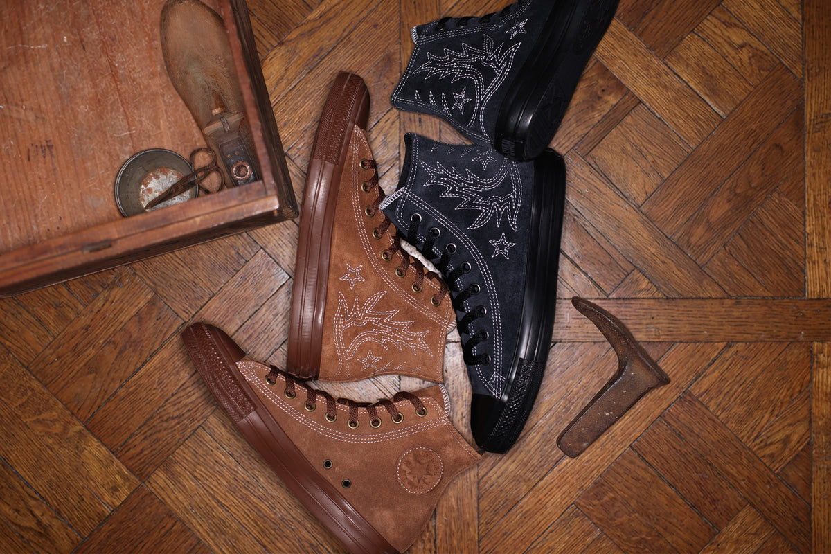 Two pairs of Converse All Star Westernstitch SU HI sneakers, one tan and one black, with white western-boot stitching and stars, laid on a worn parquet floor next to an old cobbler's box