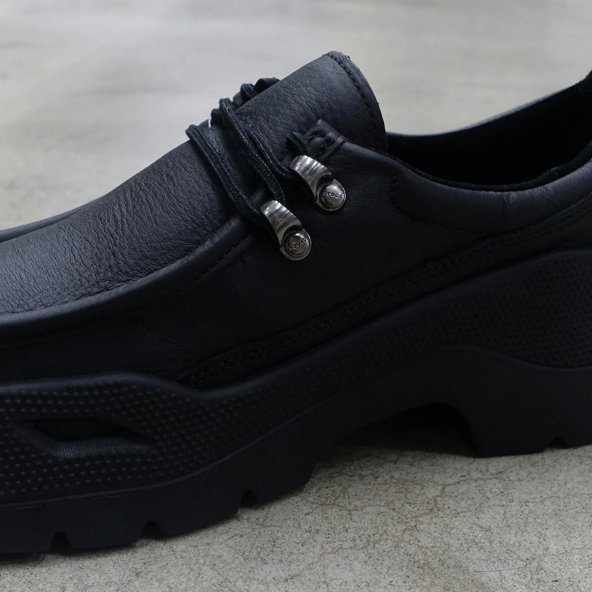 Crocs Gallery Leather Shoe in black, close-up of the leather upper and metal hiking eyelets on a concrete floor. Photo via Mita Sneakers / Highsnobiety