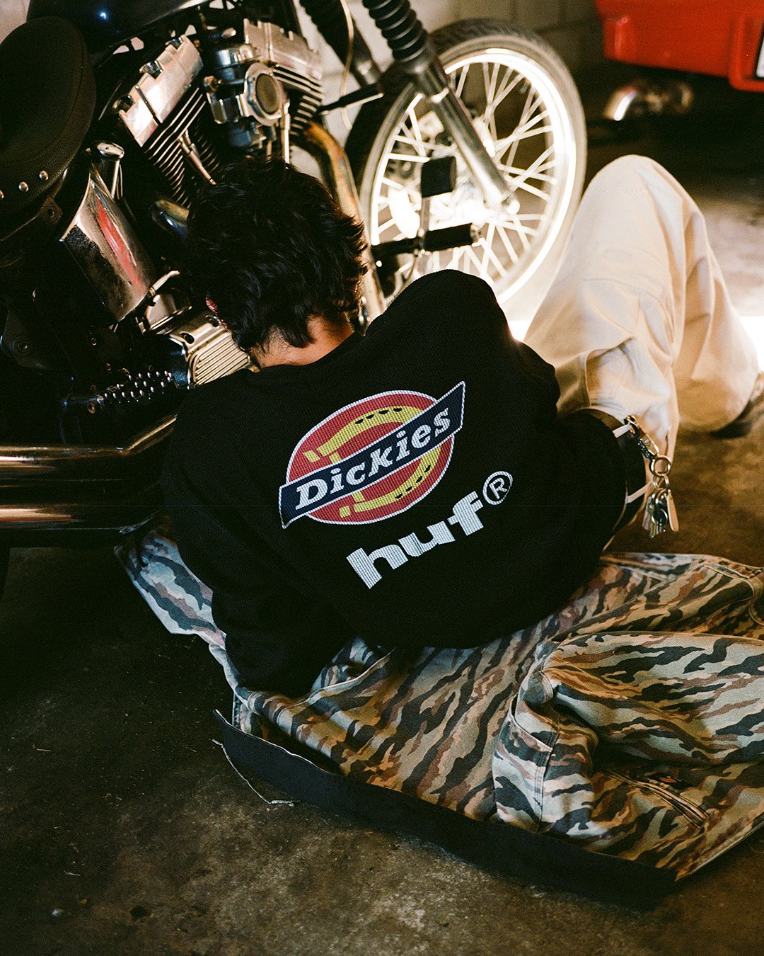 A person in a black top with the Dickies horseshoe logo stacked over the huf wordmark on the back, working on a motorcycle in a garage, from HUF Japan's official huf x Dickies teaser