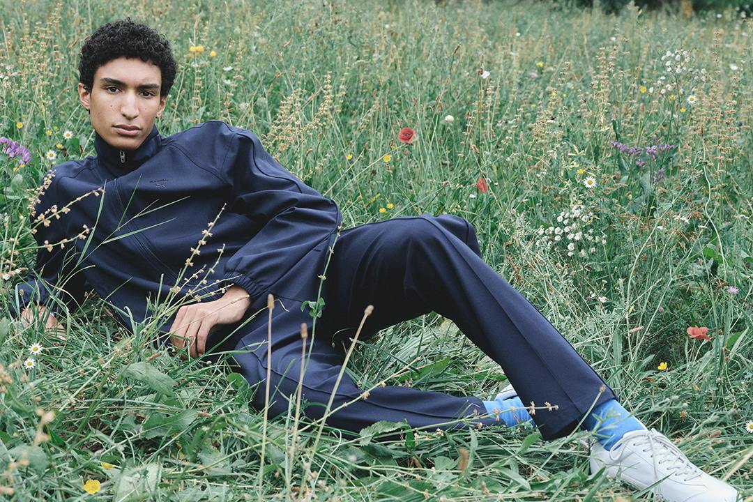 Jil Sander x PUMA T7 tracksuit in navy with white sneakers, model lying in a meadow. Photo via Highsnobiety