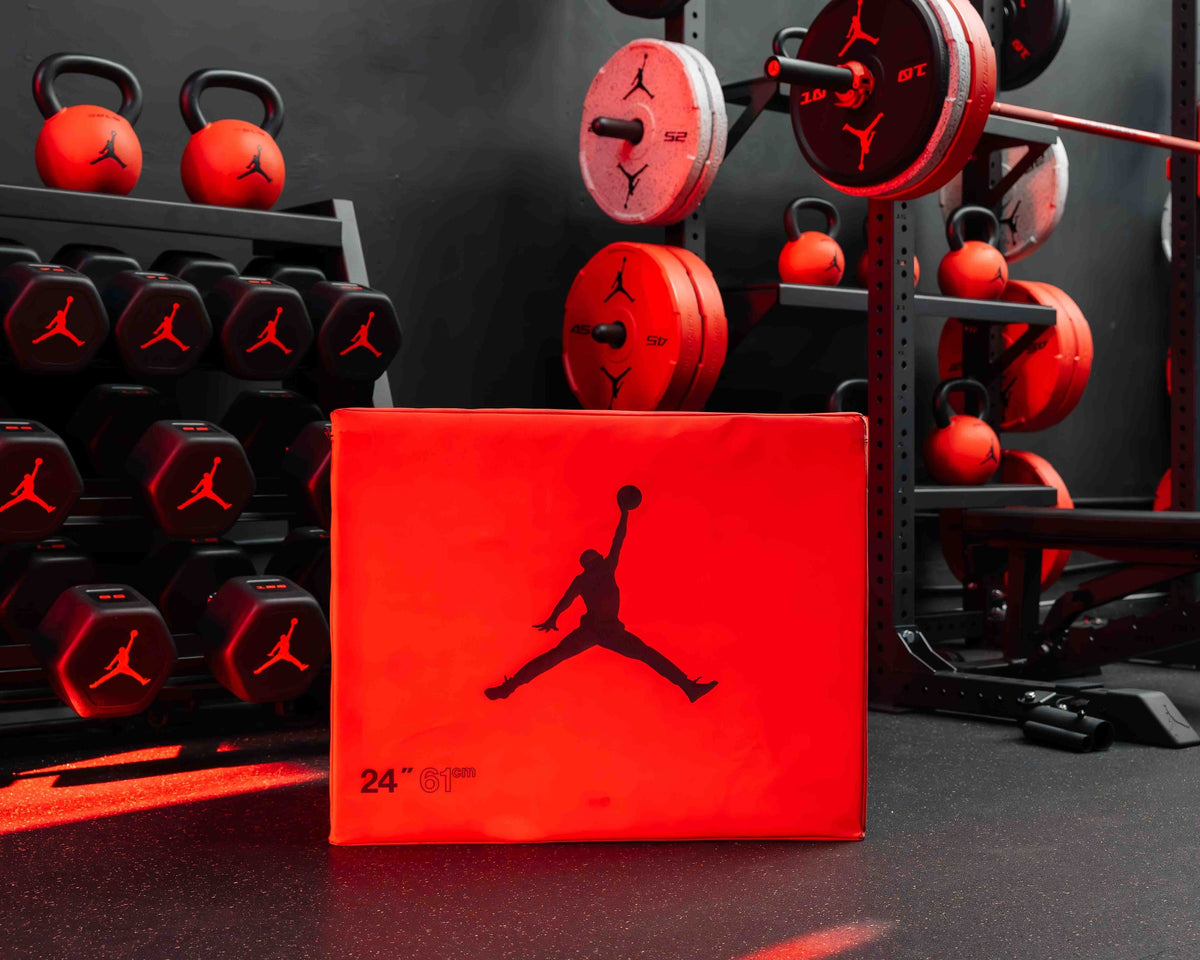 Jordan Strength soft plyo box in red with the Jumpman logo, in front of Jordan Strength dumbbells, kettlebells and bumper plates in a dark gym — official Jordan Brand image