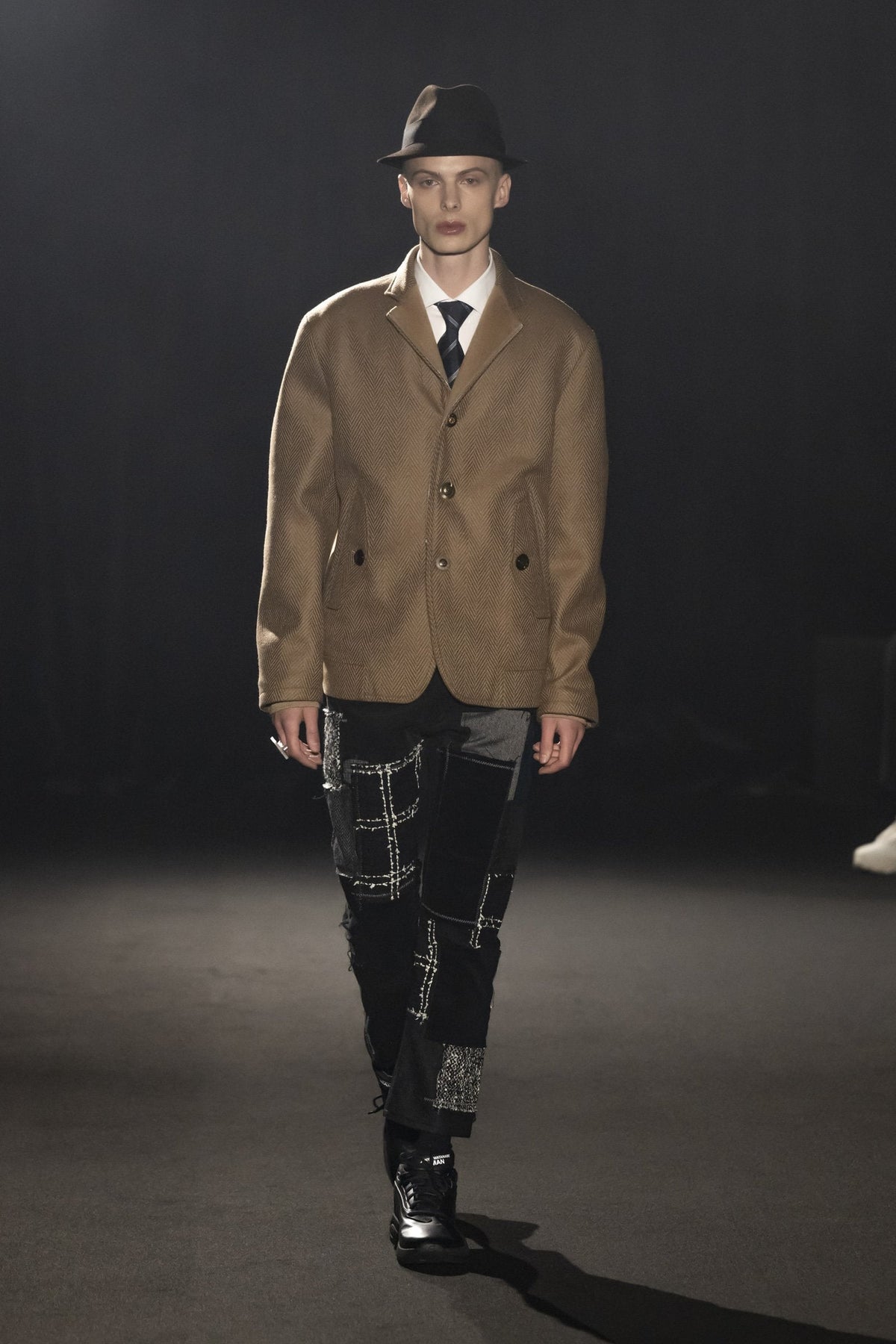 Model on the JUNYA WATANABE MAN FW26 runway in a camel coat and patchwork trousers wearing the all-black Junya Watanabe MAN x New Balance P400