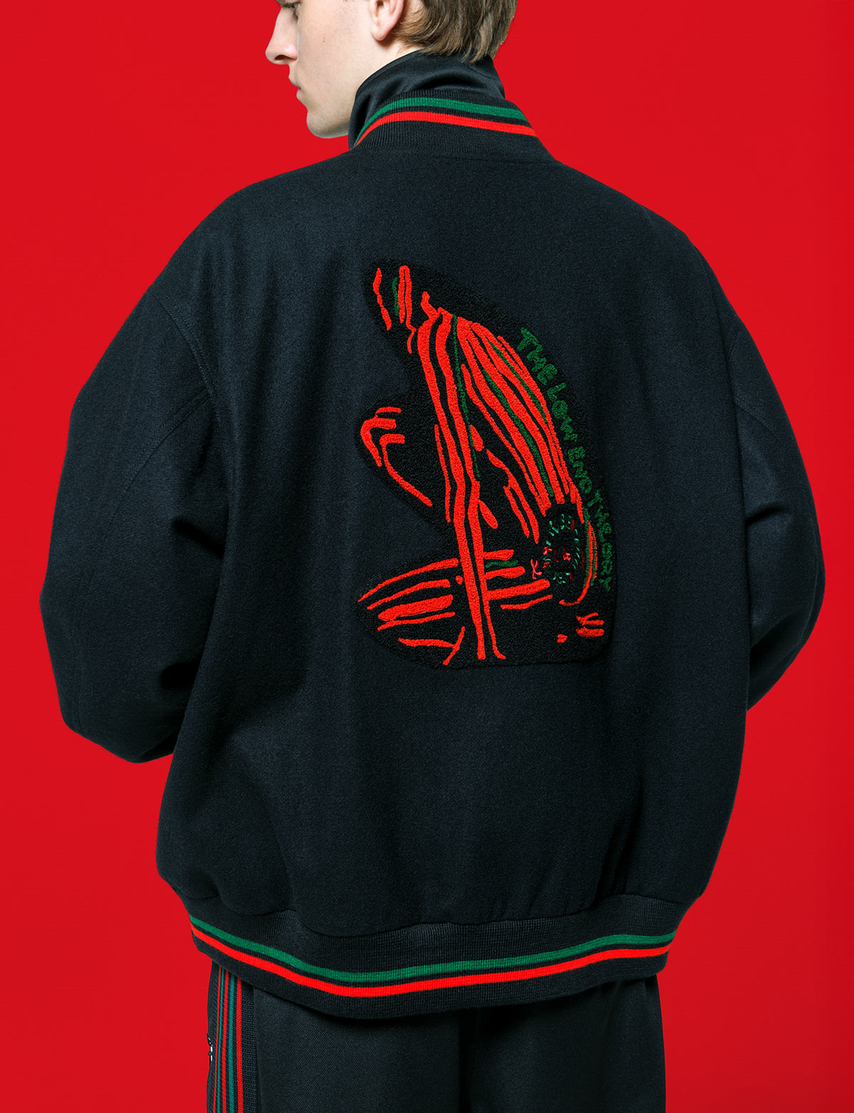 Back of the black NEEDLES x A Tribe Called Quest B.B. Jacket with a chenille The Low End Theory cover patch and red and green rib trim, against a red backdrop