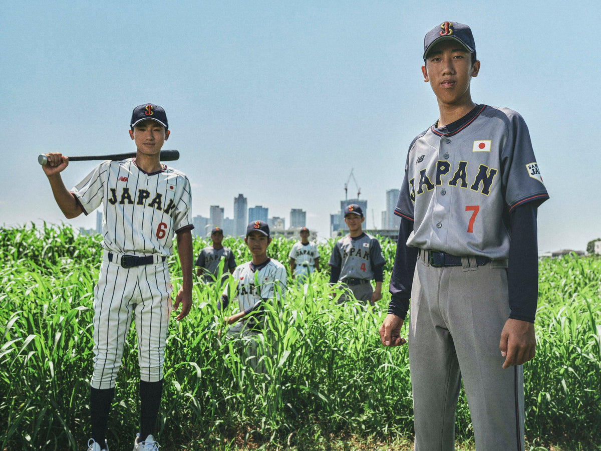 Young Samurai Japan players in the new New Balance pinstriped home and gray visitor uniforms and SJ caps stand in tall green grass with the Tokyo skyline behind them