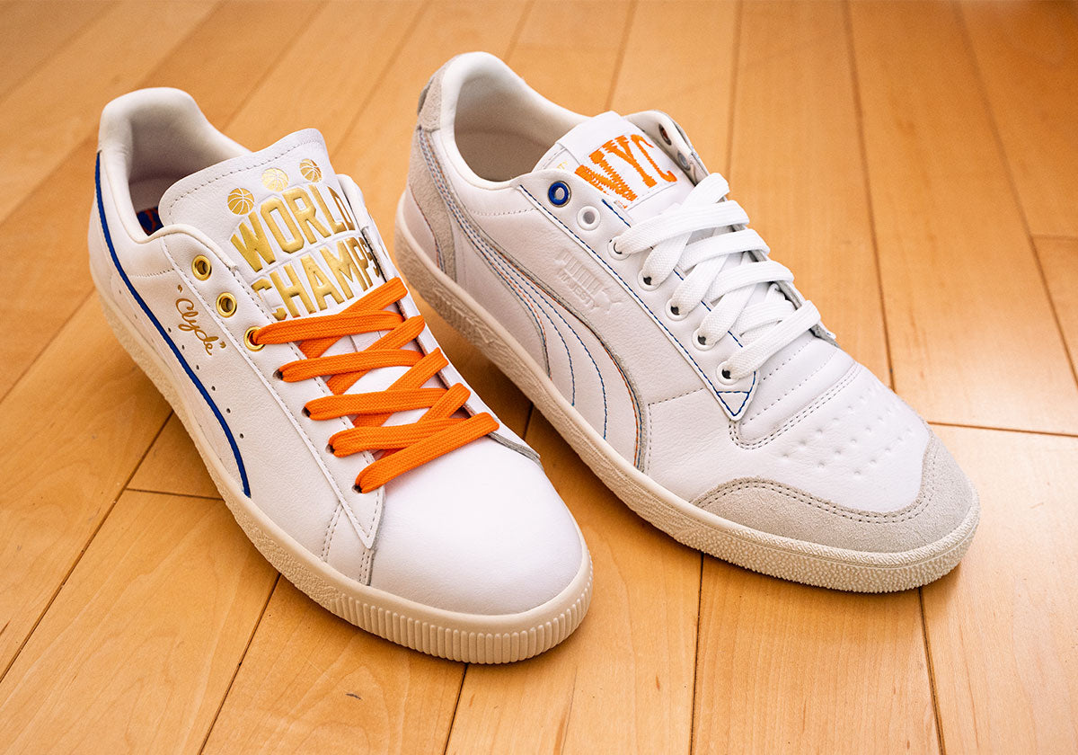 PUMA Clyde Ceremony with gold World Champs tongue and orange laces beside the PUMA Majesty Ceremony with an orange NYC tongue, on a hardwood floor