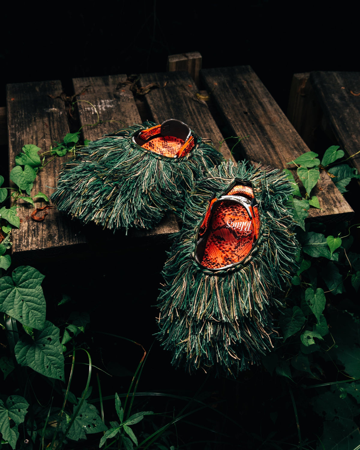 Two Sneaker Politics x Crocs Echo II Watch Your Step clogs covered in a green grass-like ghillie shroud on a wooden bench in the woods — official Sneaker Politics campaign image