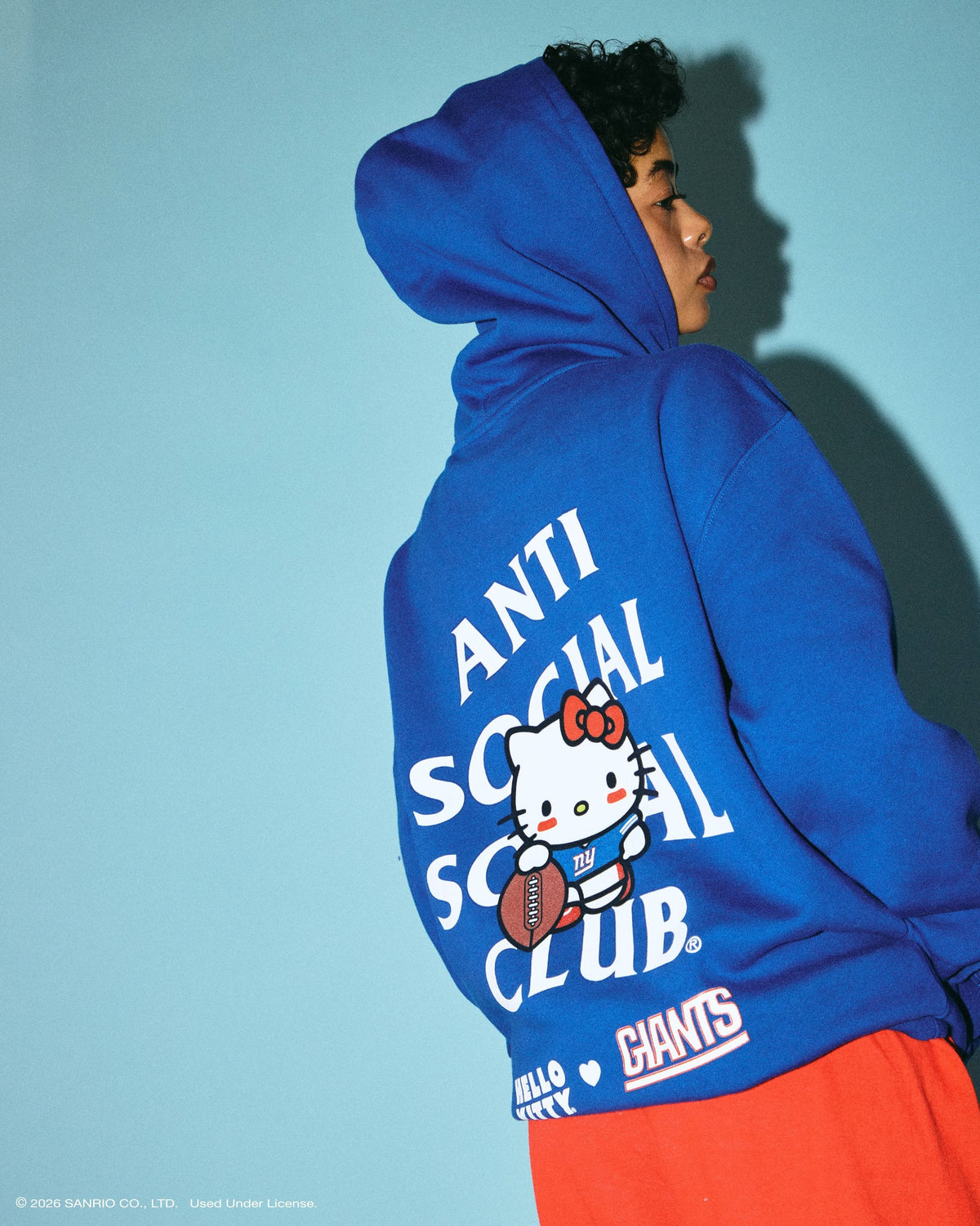 Anti Social Social Club × Hello Kitty NFL collection: model in a royal-blue New York Giants hoodie with Hello Kitty holding a football inside the ASSC logo, lookbook photo via House of Heat
