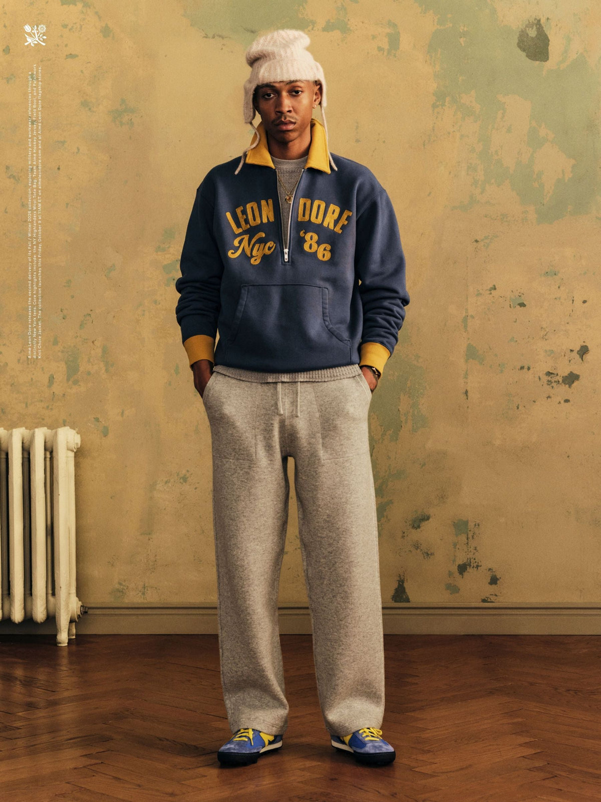 Aimé Leon Dore FW26 Delivery 2 lookbook: model in a cream beanie, navy Leon Dore NYC '86 zip sweatshirt and grey trousers wearing the blue-and-yellow Aimé Leon Dore x New Balance Gator