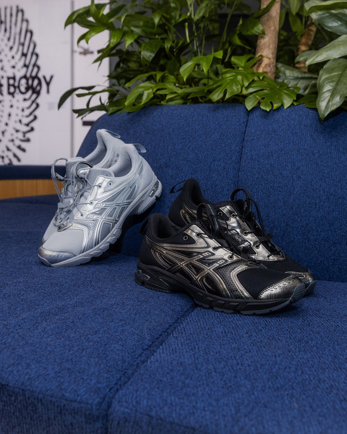 ASICS GEL-DS TRAINER LOOP in Piedmont Grey and Pure Silver and in Black and Gunmetal, sitting on a blue sofa in front of plants in the official ASICS campaign