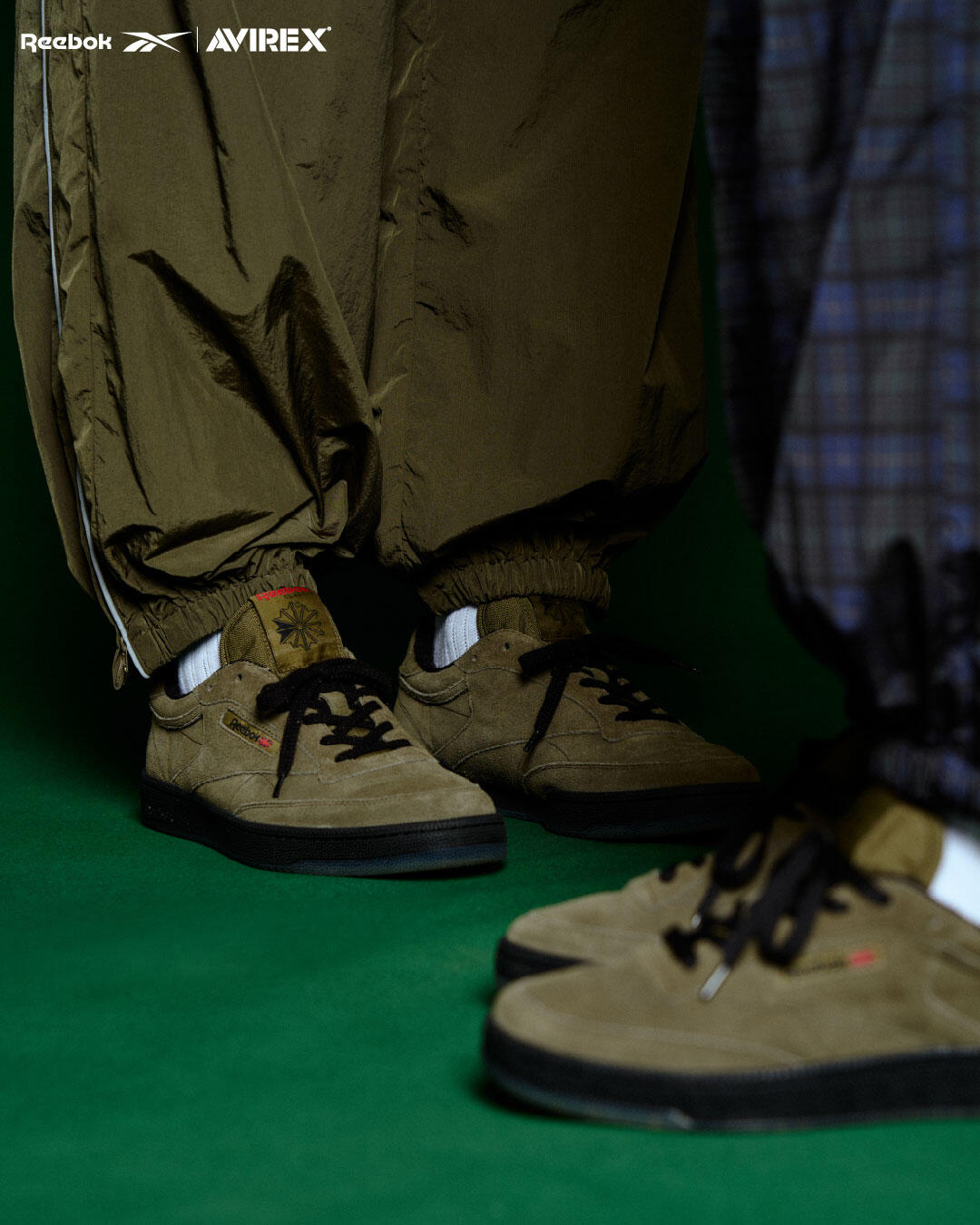 Two people in olive and plaid AVIREX x Reebok training pants wearing olive suede AVIREX x Reebok Club C sneakers on a green studio floor