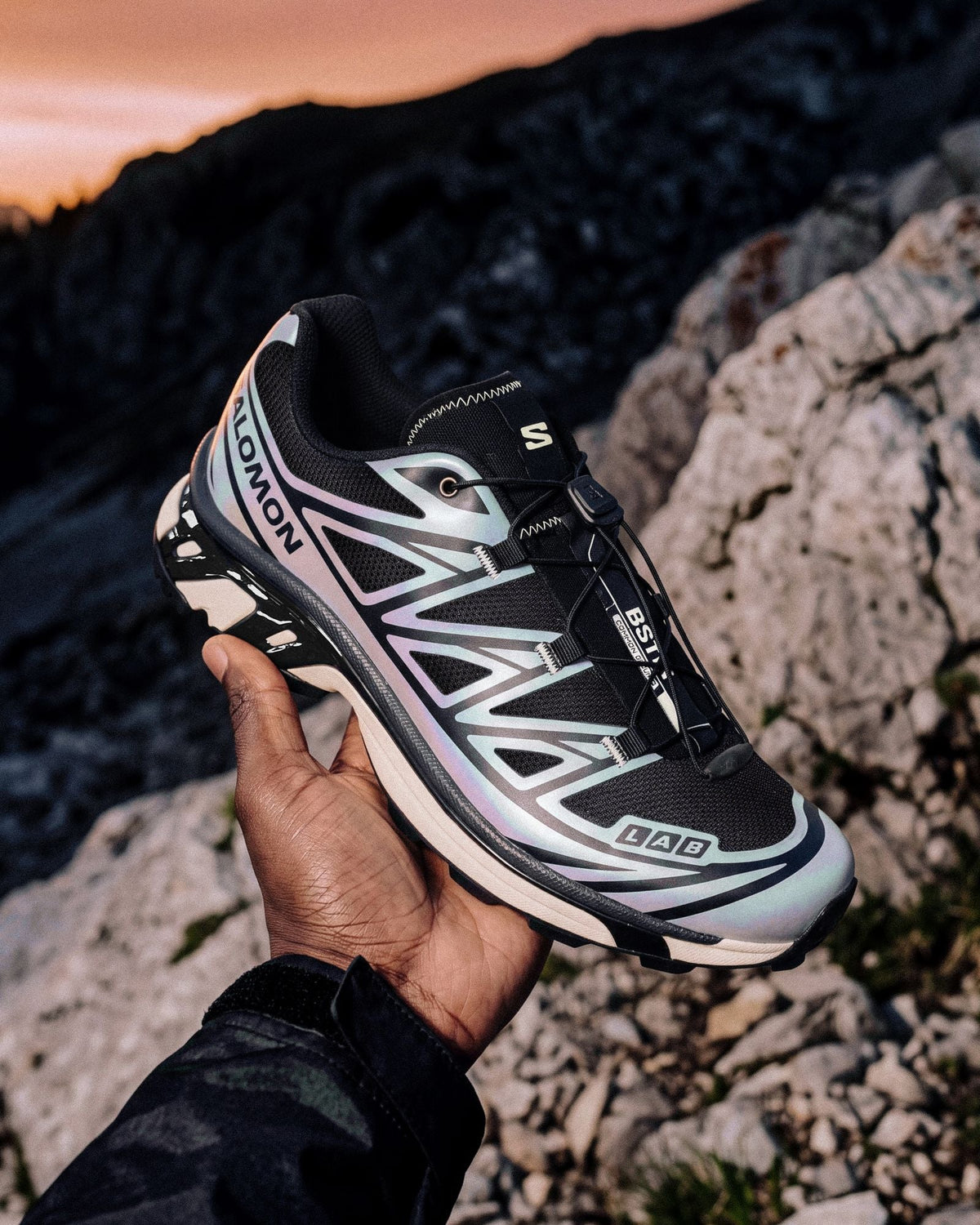 A hand holds up the BSTN x Salomon XT-6 5 AM with its iridescent silver upper against a sunrise over rocky mountains — official BSTN campaign image