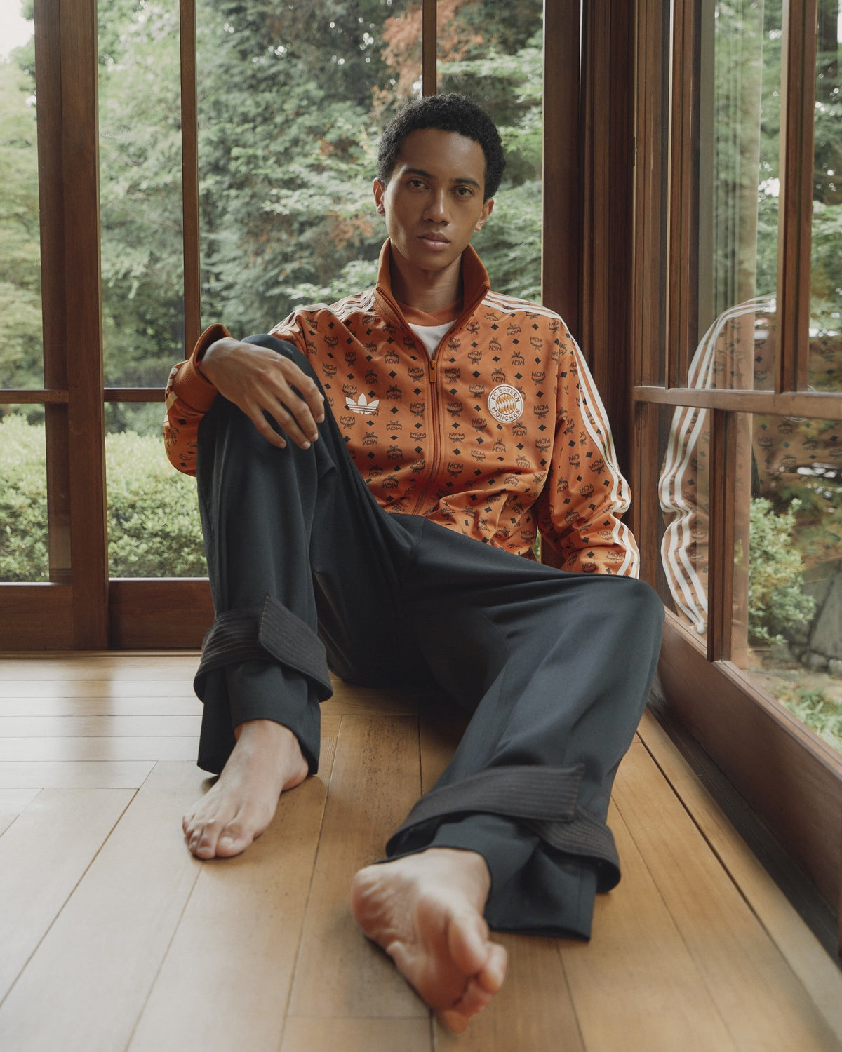 adidas x FC Bayern x MCM capsule campaign: model seated by a window in the Cognac Visetos monogram adidas track jacket — courtesy of MCM
