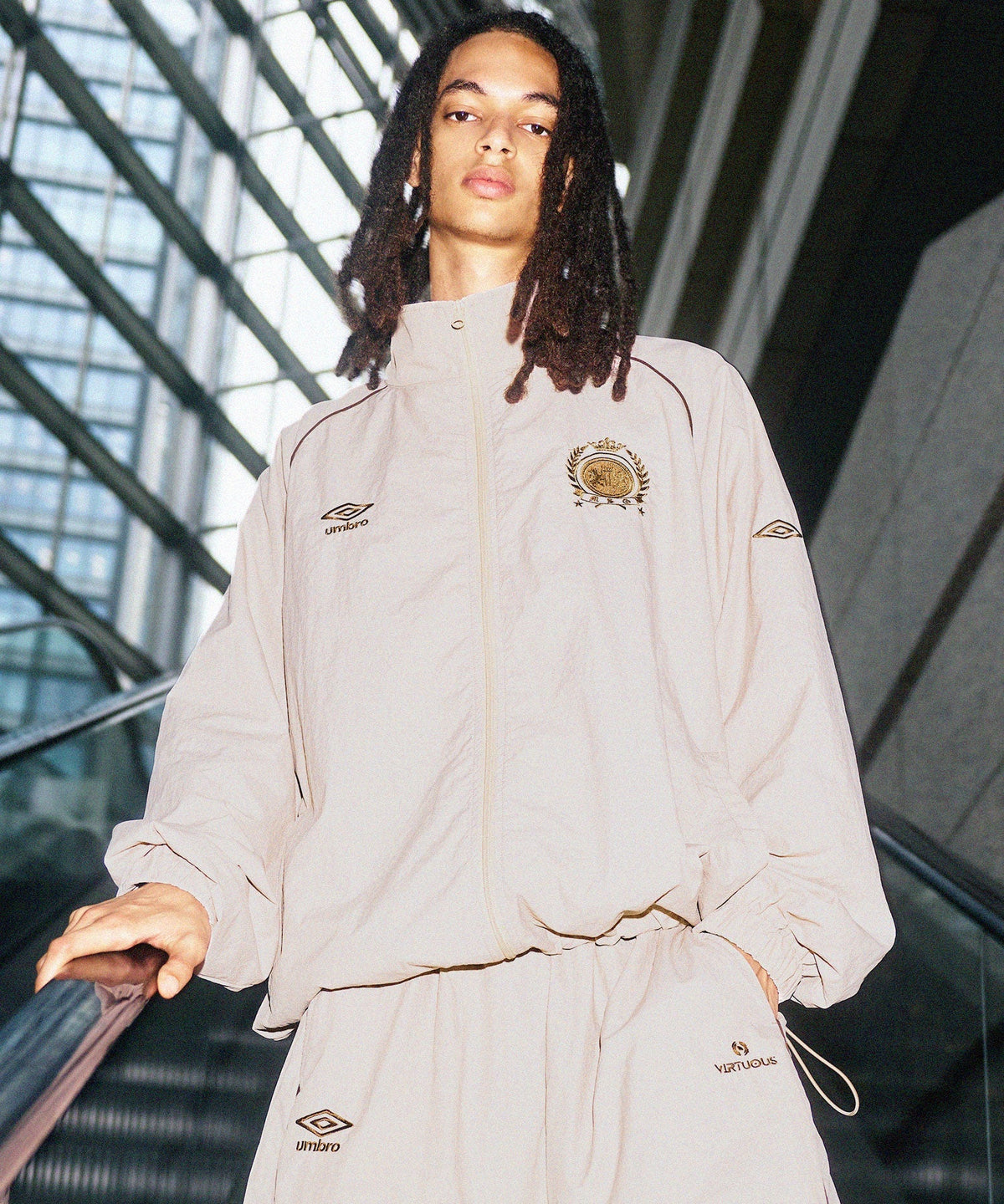 A model in the sand-beige MSG x UMBRO nylon track jacket and pants with a gold crest, riding an escalator under a glass roof