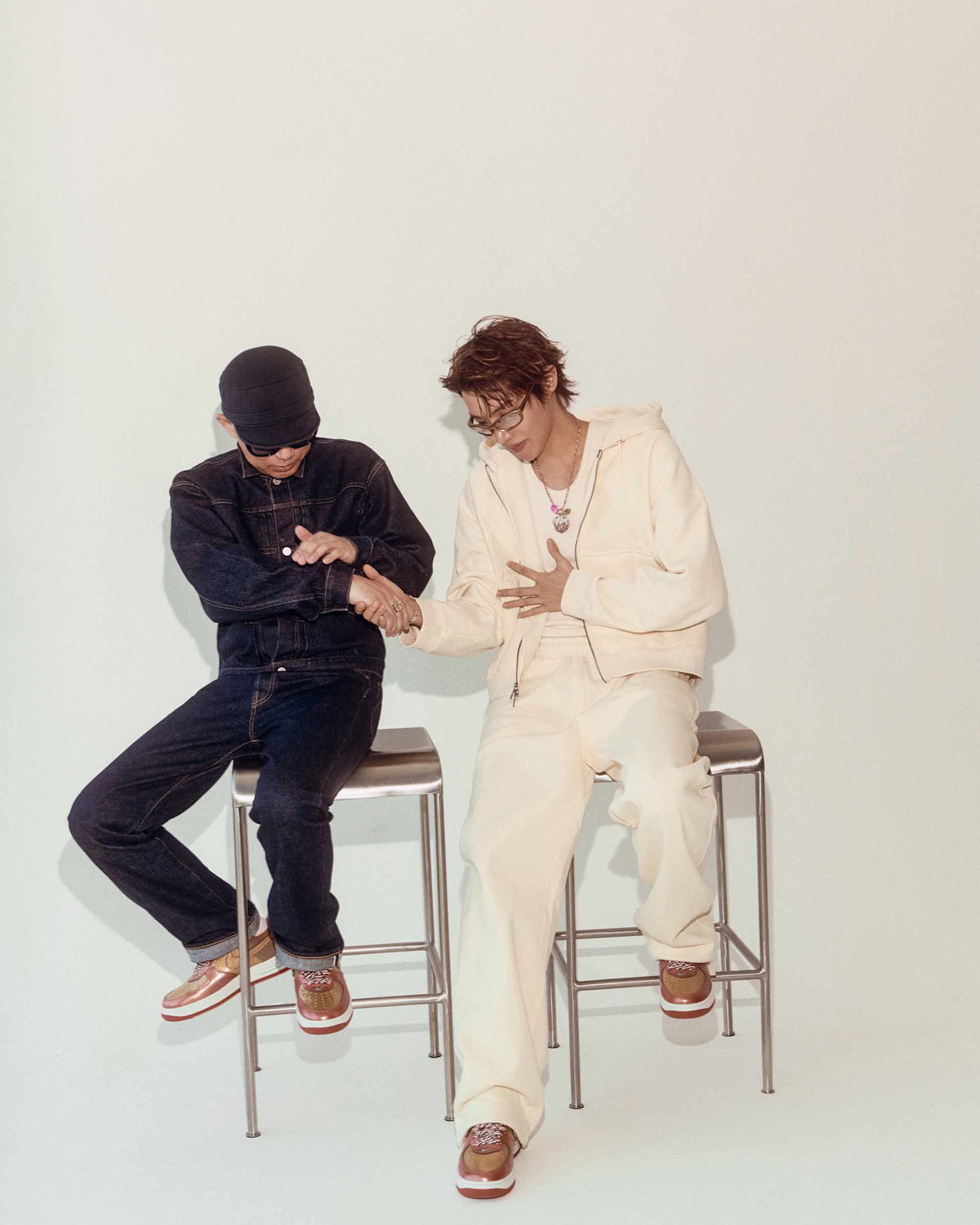 NIGO and j-hope shake hands while seated on metal stools, both wearing the Nike Air Force 1 Burnt Sunrise collaboration sneakers