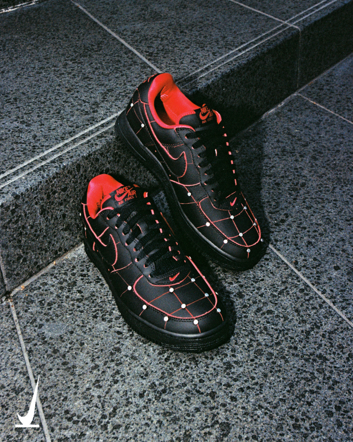 A pair of Nike Air Force 1 '01 Nightmare sneakers in black leather with Gym Red seams and silver pins, sitting on granite steps, from Nike SNKRS