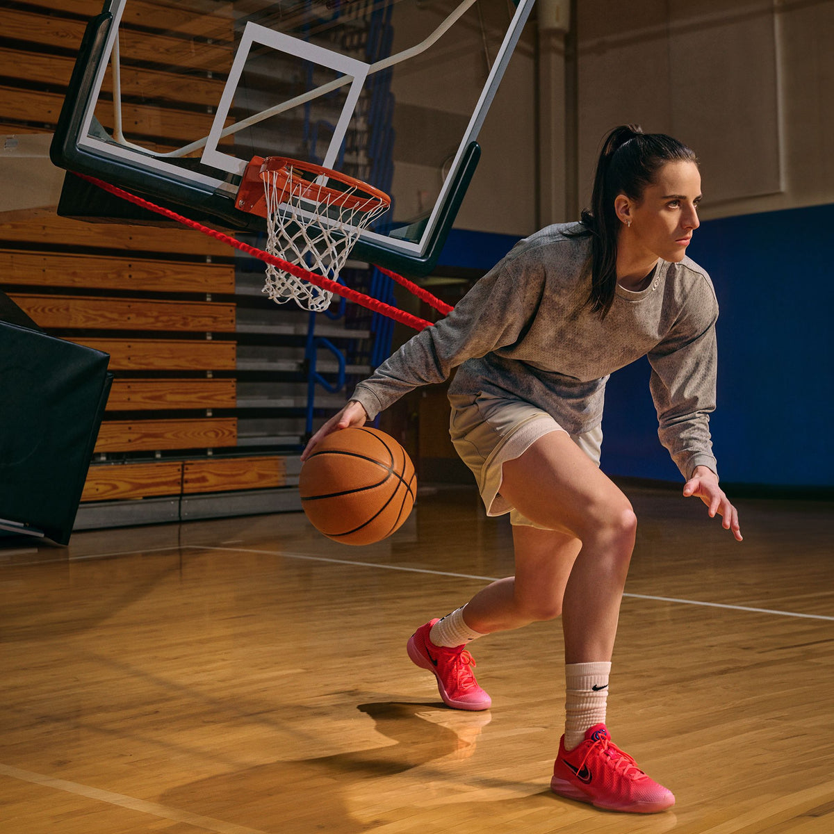 Caitlin Clark dribbling on a hardwood gym court in the hot pink Nike Caitlin 1 Midnight Fever, with a basketball hoop behind her. Official Nike image via House of Heat