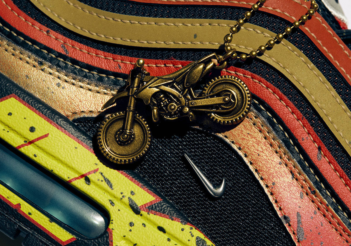 Close-up of the Doernbecher Freestyle 22 Nike Air Max 1/97 by Owen William Flint, with a brass dirt bike keychain hanging over mud-splattered red, orange and yellow wavy panels. Image via Sneaker News