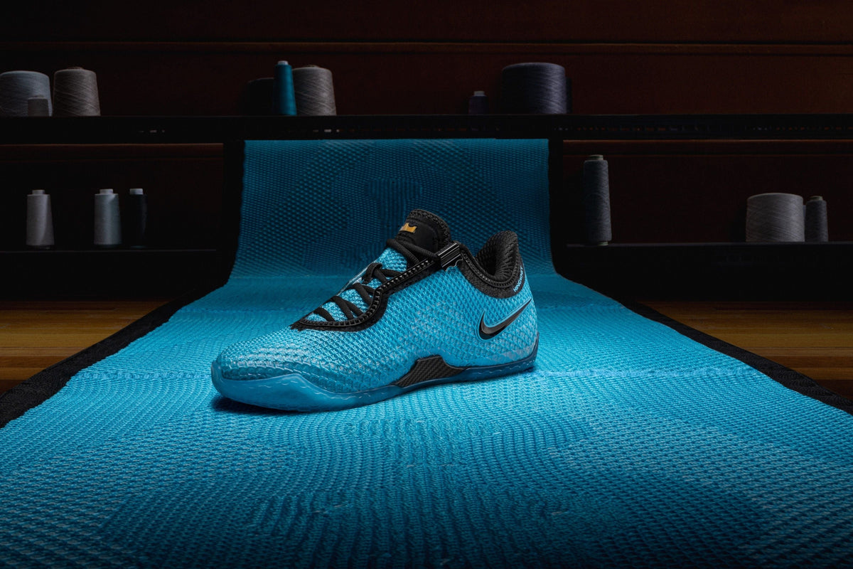 The Nike LeBron 24 in bright blue KingKnit with black laces and heel collar, sitting on a long roll of matching blue knit in a dark room lined with thread spools — official Nike image