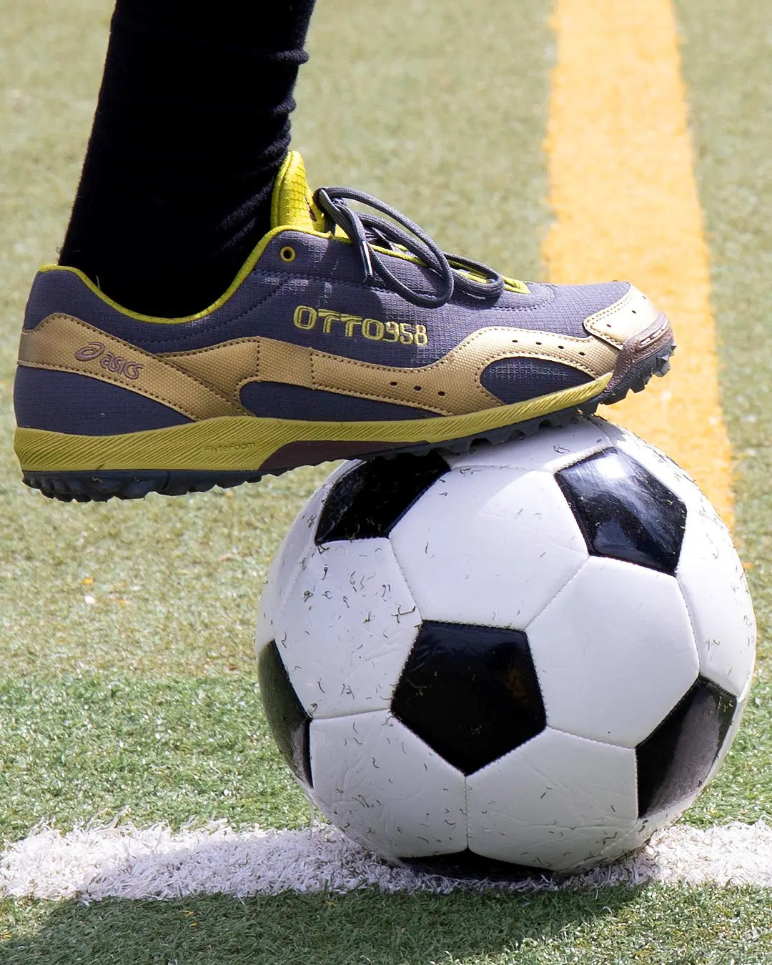 OTTO 958 x ASICS GEL-BELARRAOTTO in Obsidian Grey / Kelp, worn with the detachable gaiter, foot resting on a soccer ball on a turf pitch. Official image via OTTO 958