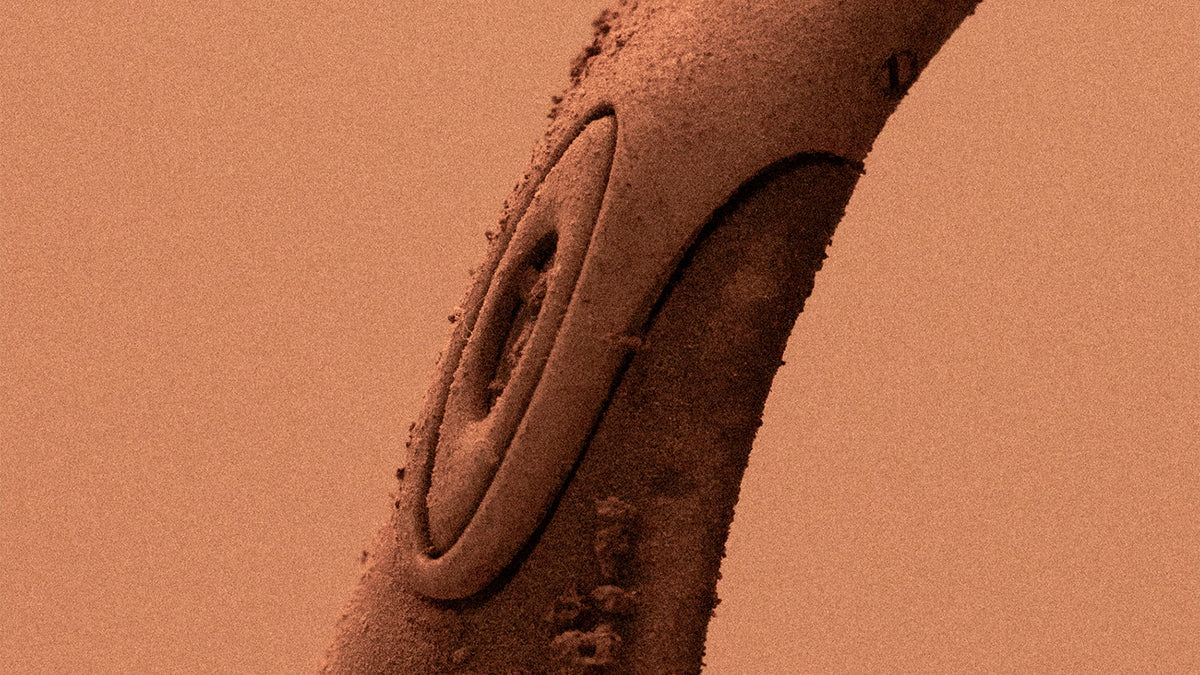 Close-up of a SATISFY x Oakley Scar temple dusted in sand against a warm orange backdrop, from the official Equipment for Our World Chapter VIII campaign