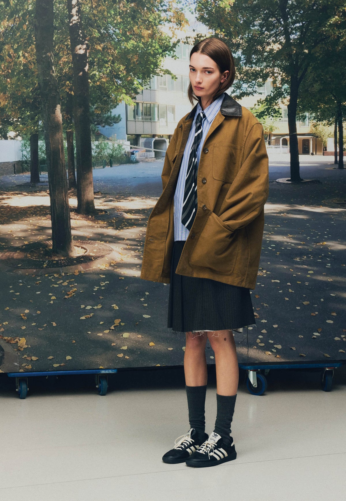 A model in a brown chore coat, striped shirt and tie wearing the black Song for the Mute x adidas Samba OG SFTM-010, from Song for the Mute's official 26.2 Playground lookbook