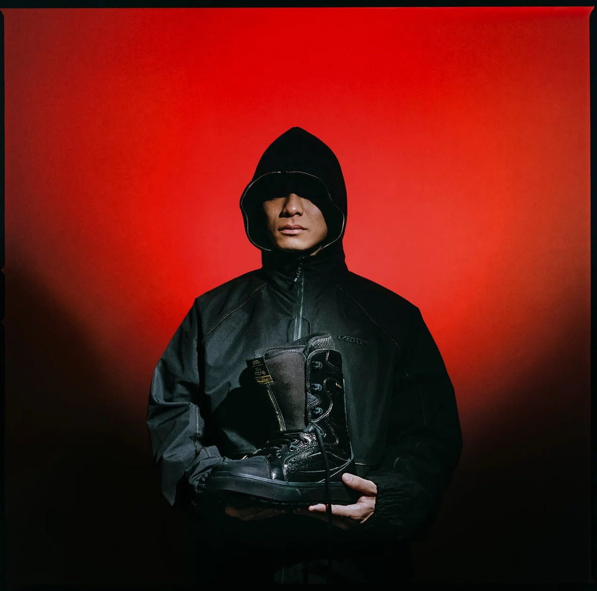 Pro snowboarder Ryo Aizawa in a black hooded jacket holds the Vans Super Standard snowboard boot against a red backdrop
