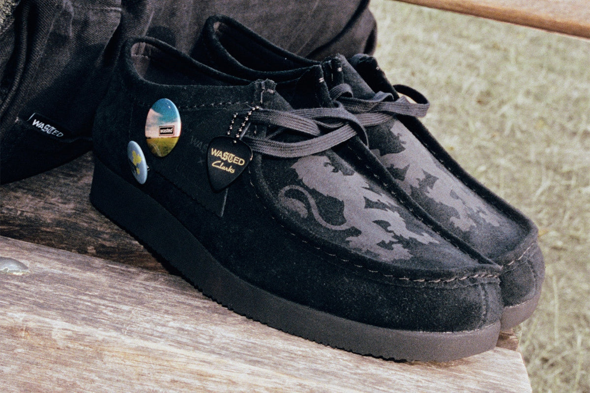 Wasted Paris x Clarks Originals Wallabee Lite in black suede on a wooden bench outdoors, with Britpop pins, a guitar pick and a Wasted x Clarks fob on the laces. Photo via Hypebeast